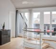 Spacious, modern room with light wooden flooring, white walls, and large windows with sheer curtains. A sleek black cabinet with a potted plant sits near the wall, and a curved white staircase with a glass railing leads upwards.