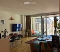 Modern living room with wooden flooring, large sliding glass doors leading to a balcony, sofa, coffee table, dining table with chairs, and a television mounted on the wall.