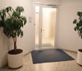 A clean, modern hallway with white walls, a polished stone floor, and a blue doormat in front of a glass door. Two large potted plants flank the entrance.