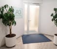 Entrance area with white walls, a glass door with a sign, two large potted plants, and a blue floor mat.