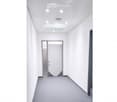 Modern, empty hallway with white walls, grey carpet, and recessed lighting. A single door is visible at the end of the corridor.