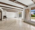 Bright, open space with exposed wooden beams, tiled floor, and large glass doors leading to an outdoor area. Features a built-in brick fireplace and white walls.