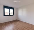 Empty room with light wooden flooring and white walls, featuring a large window with black frame and a visible outdoor view.