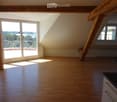 Bright, empty attic room with wooden floors, large windows, and exposed wooden beams. Features a balcony area with outdoor view.