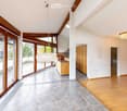Bright, open hallway with wooden floors, large windows, and modern lighting fixtures. The space features a mix of tiled and wooden flooring, leading to a kitchen area with wooden cabinets and a countertop.