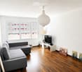 Modern living room with wooden flooring, large windows, grey sectional sofa, coffee table, TV, and decorative artwork.