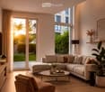 Modern living room with a large sectional sofa, coffee table, armchair, floor lamp, and large windows with sheer curtains. The space features a light-colored carpet, wooden flooring, and decorative plants.