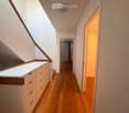 Modern hallway with wooden flooring, white walls, built-in storage unit with drawers, and a skylight providing natural light. The hallway leads to another room with a visible door and window.