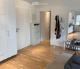 Bright living room with wooden flooring, a grey sofa, a large rug, a white wardrobe, a shoe rack, and a visible kitchen area with cabinets and appliances.
