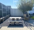 Well-maintained garden with a stone table, benches, and a barbecue, surrounded by gravel and a modern fence.