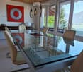 Modern dining room with a large glass table, leather chairs, wooden sideboard, potted plant, and decorative artwork. Large windows offer a scenic mountain view.