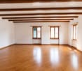 Empty room with wooden flooring, white walls, exposed wooden ceiling beams, and three windows with wooden frames and radiators beneath them.