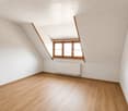 Empty attic room with wooden flooring, white walls, and a slanted ceiling. Features a single window with wooden frame and a radiator below it.