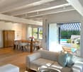 Bright living and dining area with wooden flooring, white sofa, coffee table, dining table with chairs, and a view to a patio with a swimming pool.