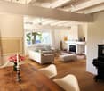 Bright living room with wooden flooring, white walls, large windows offering a view, a fireplace, a sofa, ottoman, dining table, chairs, and a piano.