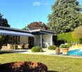 Well-maintained garden with a swimming pool, patio furniture, and a modern house with a striped awning.