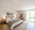 Modern bedroom with a large bed, nightstands, lamps, artwork, clothing rack, and plants.
