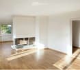Empty room with light wooden flooring, white walls, and a central fireplace. Large window on one side allows natural light to enter.