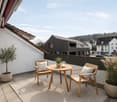 A modern balcony with a small round table, two wooden chairs, and potted plants, overlooking a town with houses and greenery.