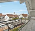 Balcony with metal railing overlooking a town with traditional houses, a church tower, and a clear blue sky.