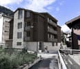 Modern multi-story building with stone and wood facade, large windows, balconies, and surrounding greenery.