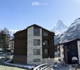 A modern stone and wood building with large windows and balconies, situated in a picturesque alpine village with snow-capped mountains in the background.