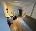 A cozy, multi-functional room with a sofa, desk, bookshelf, and guitar, featuring wooden flooring and large windows.