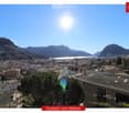 Panoramic view from a balcony showing a cityscape with buildings, a lake, and mountains under a clear blue sky.