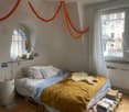 Cozy bedroom with a wooden bed, pillows, blankets, and a small table with books. Large windows with white curtains and a radiator are visible.