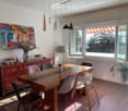Bright dining area with a long wooden table surrounded by modern chairs, a sideboard with decorative items, a lamp, and a large painting on the wall. Large windows allow natural light to fill the space.