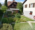 Well-maintained garden with lawn, flower beds, and a small wooden shed. The garden is bordered by a white fence and features a variety of plants and shrubs. A house with a red-tiled roof and white walls is visible in the background.
