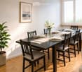 Dining room with a dark wooden table set for six, surrounded by six chairs with beige cushions. A vase with white lilies is placed at the center of the table, and wine glasses are set around. A potted plant is on the left side, and a framed picture hangs