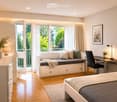 Modern bedroom with a bed, built-in sofa, desk, chair, lamp, and decorative items. Large windows and a balcony offer a view of greenery.