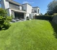 Well-maintained lawn in front of a modern house with large windows and a balcony, surrounded by greenery and a stone wall.
