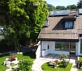 Modern two-story house with solar panels on the roof, large windows, and a well-maintained garden with a stone pathway and various plants.