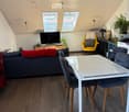 Modern living room with wooden flooring, a grey sofa, a white dining table with four chairs, a yellow armchair, a television, and several plants.