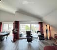 Bright living room with wooden flooring, large windows, and a balcony. Features a sofa, chairs, a table, a fireplace, and decorative statues.