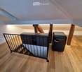 An attic space with wooden flooring, exposed wooden beams, and a staircase with a safety railing. A small black cabinet and a black mini-fridge are present.
