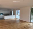 Open-plan living and kitchen area with light wood flooring, white cabinetry, and large windows leading to a balcony.