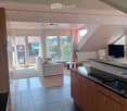 Open-plan living and kitchen area with wooden beams, large windows, and a balcony. Features a sofa, coffee table, television, and modern kitchen with cabinets and a sink.