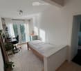 Bright bedroom with a wooden bed, desk, chair, laptop, and a plant near the window. The room features white walls, wooden flooring, and large windows with blinds.