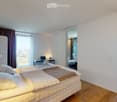 Modern bedroom with a large bed, wooden flooring, a desk with a laptop, a small table with a plant, and a mirror on the wall. The room has large windows with curtains and a view of the city.
