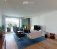Modern living room with wooden flooring, large sliding glass doors leading to a balcony, a blue rug, a white armchair, a black leather chair, a small wooden side table, a television, and a lamp.