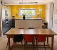 Bright kitchen with a yellow accent wall, wooden dining table, and modern appliances. Features include a bar counter, stools, and a potted plant on the counter.