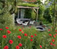 A lush garden with red poppies, a wooden pergola, white chairs, and a table, surrounded by greenery and trees.