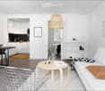 Modern, minimalist living room with white walls, light wood flooring, and a white sofa with a brown throw pillow. A round wooden coffee table holds a candle and a vase with dried flowers. A metal wire chair is near the coffee table. The room features a sm