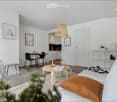 Modern living room with white walls, a white sofa with orange pillow, a round wooden coffee table, a metal wire chair, a small dining table with black chairs, a shelf with decorative items, and a hanging woven lamp.