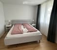 Bedroom with a double bed, nightstands, lamps, and curtains. The bed has a grey headboard and is made up with pink bedding.