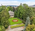 Aerial view of a lush garden area with a circular flower bed, a fountain, a house, and surrounding trees, with a cityscape in the background.