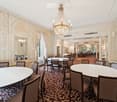 Elegant dining area with round tables, chairs, ornate chandelier, large windows with curtains, and a patterned carpet.